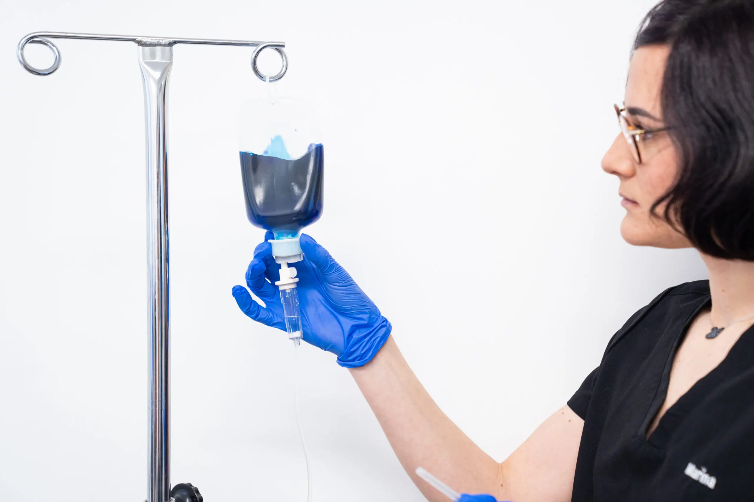 A healthcare worker wearing blue gloves holds a transparent drip bag filled with methylene blue liquid, attached to a drip stand, against a white background.