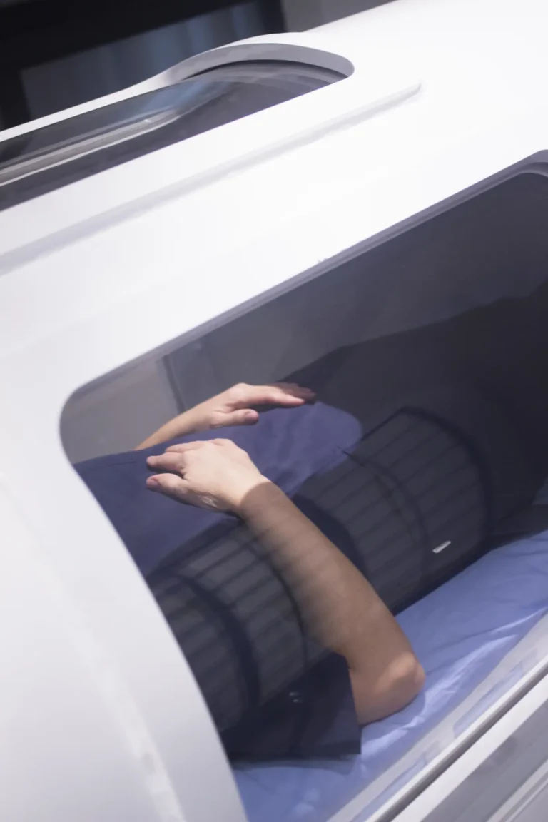 A person lies inside a Hyperbaric Oxygen Therapy machine, with their hands folded on their abdomen and visible through a window.