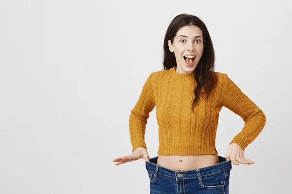 Woman in a mustard jumper holding out oversized jeans, smiling with a surprised expression—showcasing the impressive results of NAD+ Weight Loss against a plain background.