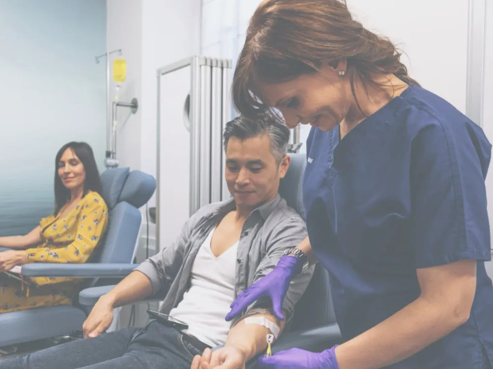 A nurse takes blood from a seated man while another woman waits in the background, both in a medical clinic where IV Booster treatments and antioxidant therapies are offered.