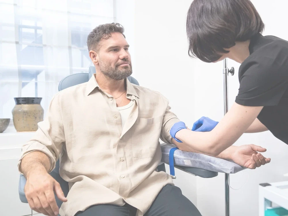A man sits in a medical chair whilst a healthcare professional wearing gloves prepares to take blood from his arm using a needle and tourniquet, possibly as part of a CRT cell-rejuvenation therapy session.