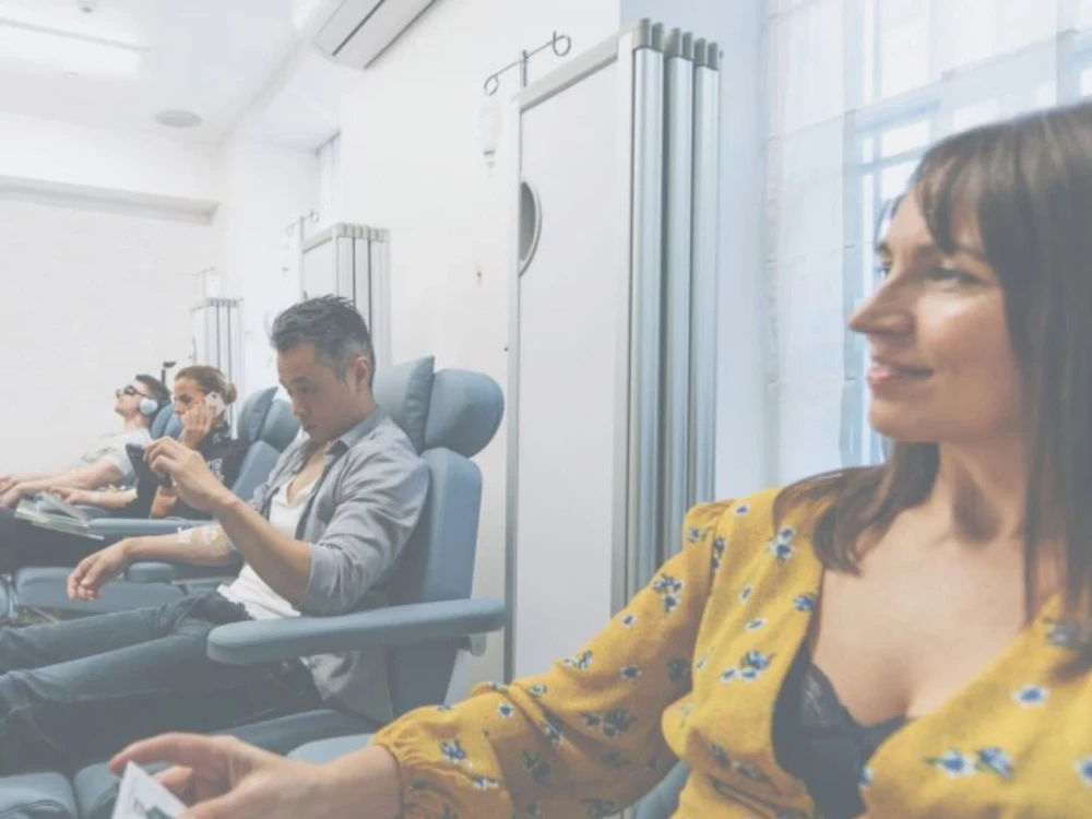 Four adults sit in reclining chairs in a bright, clinical room, each appearing relaxed and engaged in individual activities during their IV therapy sessions.