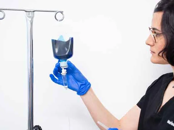 A person wearing blue gloves adjusts a Methylene Blue Infusion drip bag on a metal drip stand against a plain white background.