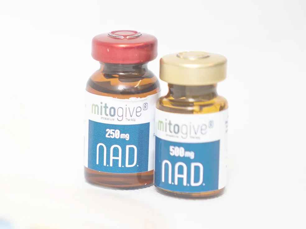 Two glass vials labelled mitogive N.A.D., one with 250 mg and a red cap, the other with 500 mg and a gold cap, contain a yellowish liquid used in NAD+ Anti-Ageing IV Therapy.