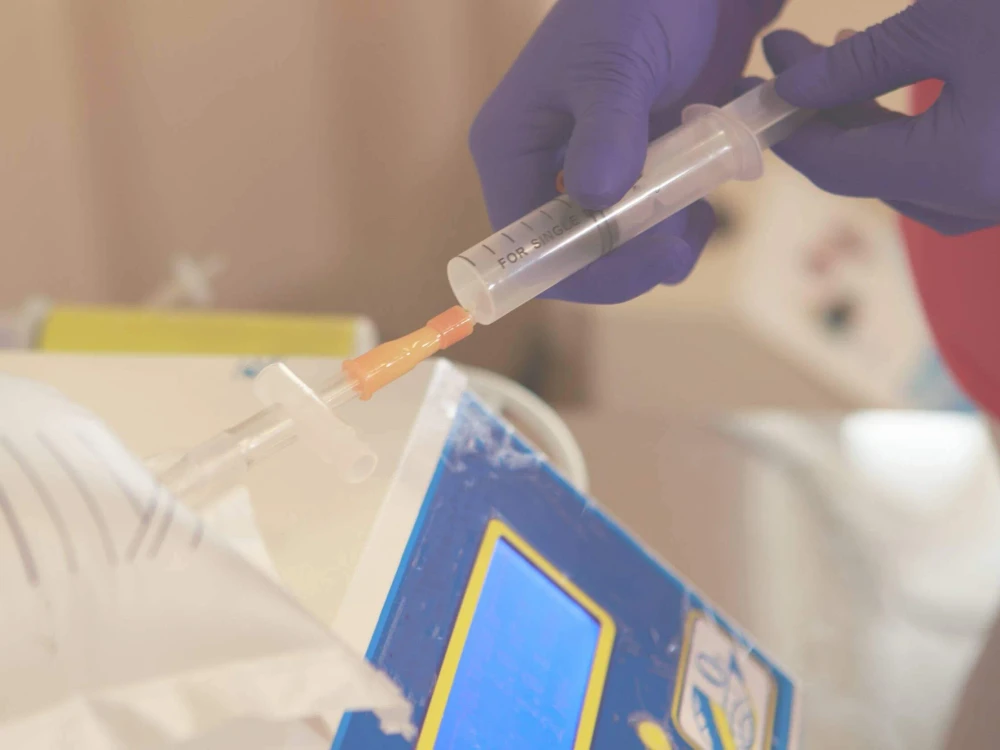 A person wearing purple gloves injects liquid into a medical tube using a syringe near a blue and yellow device, preparing for ozone therapy.