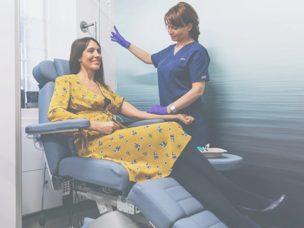 A woman in a yellow dress sits in a medical chair whilst a healthcare professional in scrubs and gloves prepares her for a Skin Brightening IV procedure.