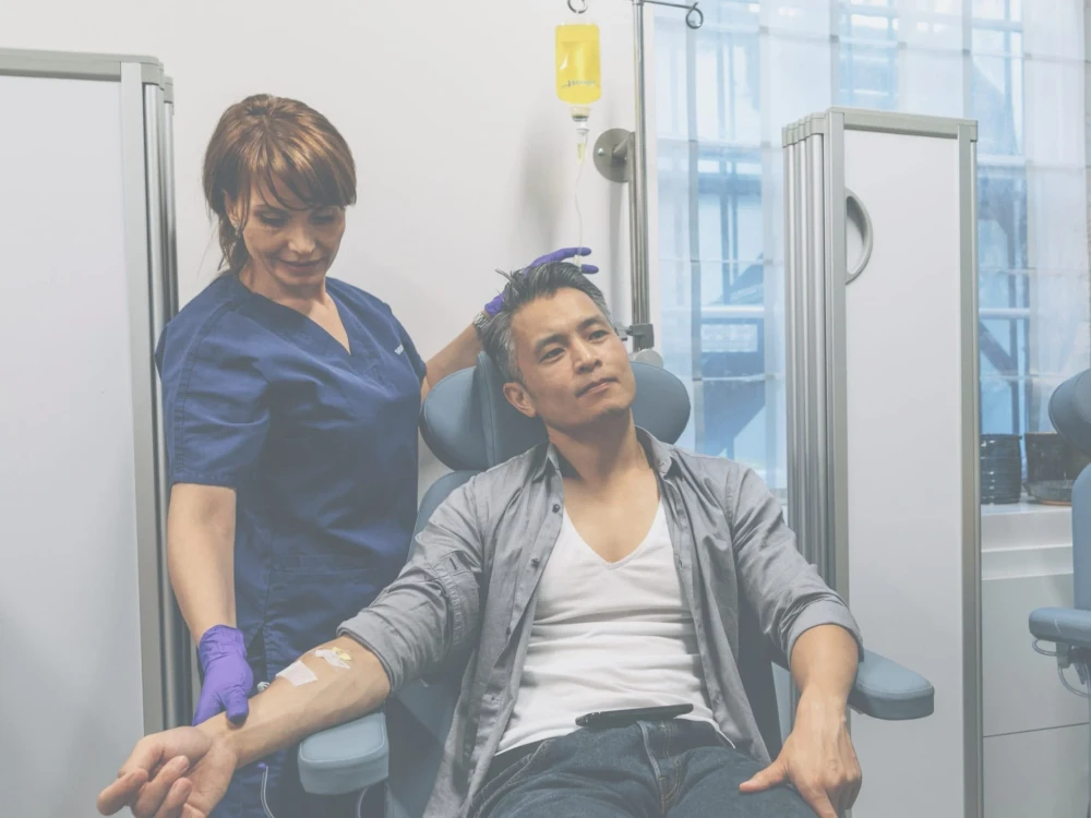 A nurse in blue scrubs assists a man receiving an IV therapy drip whilst seated in a medical clinic.