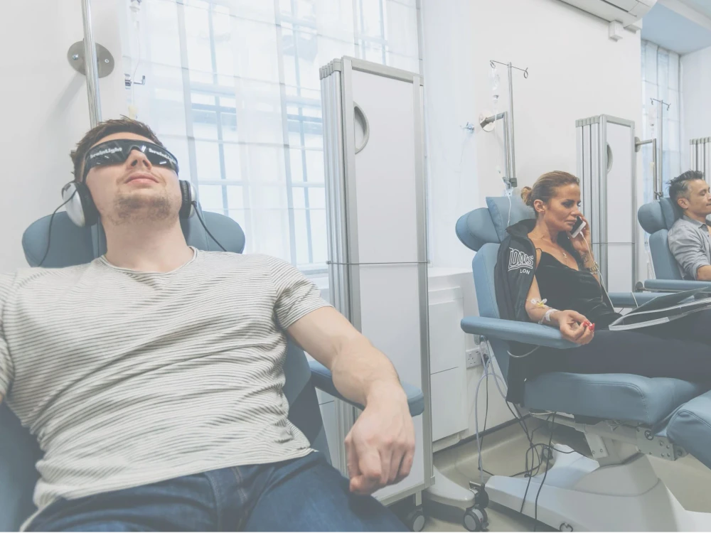 Three people sit in reclining chairs receiving IV Therapy; one man wears headphones and dark glasses, a woman uses a laptop, whilst another man relaxes in the background, all experiencing an Energy IV Booster for a revitalising energy boost.