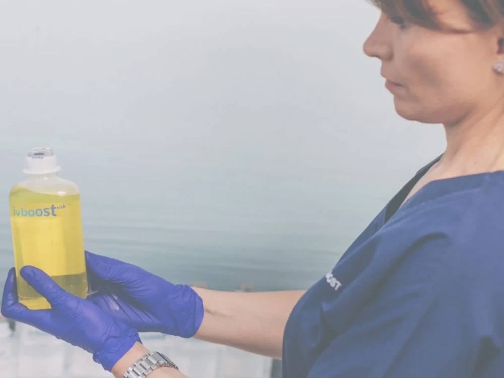 A person in blue medical scrubs and gloves holds a clear bottle with yellow liquid labelled ivboost.uk, highlighting the use of High-Dose Vitamin C for intravenous therapy.