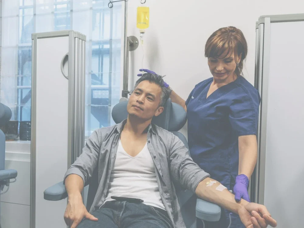 A nurse in blue scrubs assists a seated man receiving a vitamin infusion through intravenous therapy in a medical clinic setting.