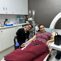 A woman lies on a medical bed covered with a red blanket, with a healthcare professional beside her in a clinical setting. Medical equipment and supplies are visible in the background.