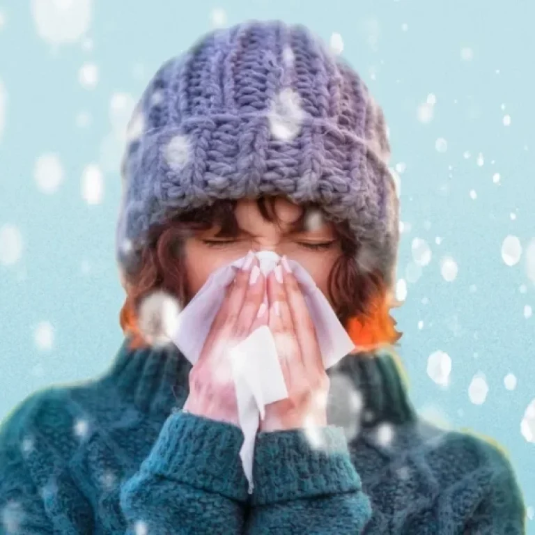 A person wearing a knitted hat and jumper blows their nose with a tissue while standing in falling snow, highlighting the importance of winter health and ways to boost immunity during the colder months.