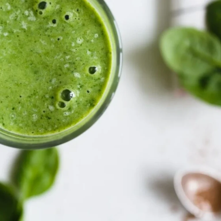 A close-up of a glass filled with a green smoothie, with fresh spinach leaves and a spoon with powder in the background—perfect for those seeking tips for better nutrition.