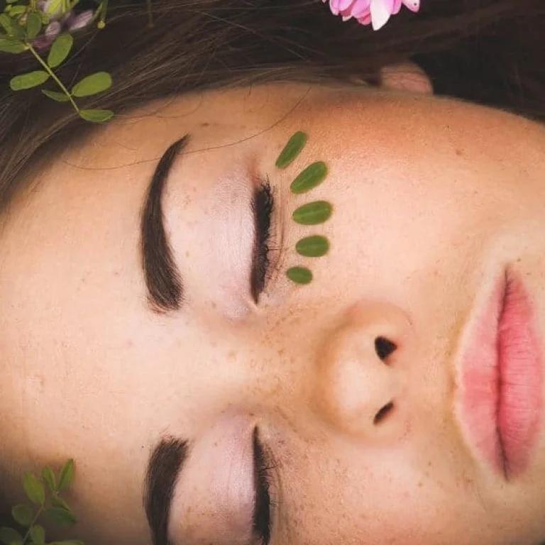 A close-up of a person with closed eyes, freckles, and green leaf petals under one eye. Pink flowers and green leaves frame their face, highlighting the natural beauty of beautiful skin.