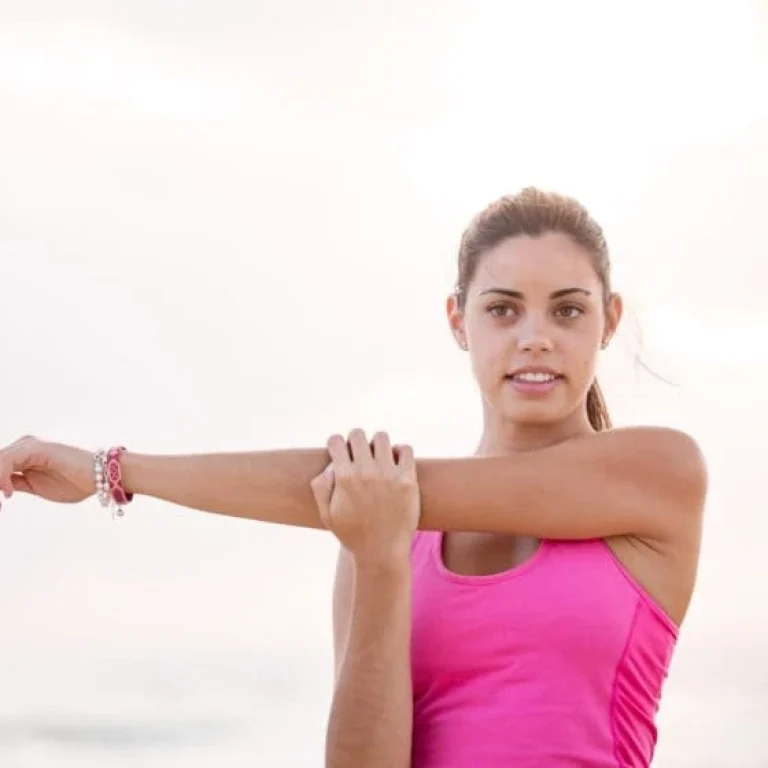 A woman in a pink vest top stretches her arm across her chest outdoors, looking towards the camera—embracing wellbeing much like exploring the 7 Major Benefits of NAD+ IV Therapy.