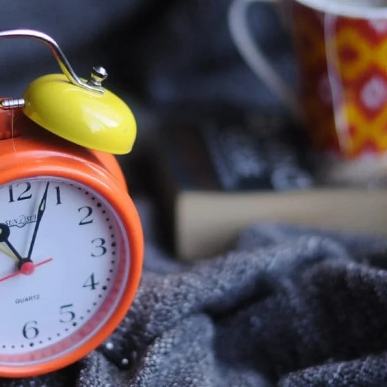 An orange and yellow analogue alarm clock shows 8:00, placed on a grey fabric with a book and a red patterned mug in the blurred background—perfect for pondering the best time for your vitamin IV drip.