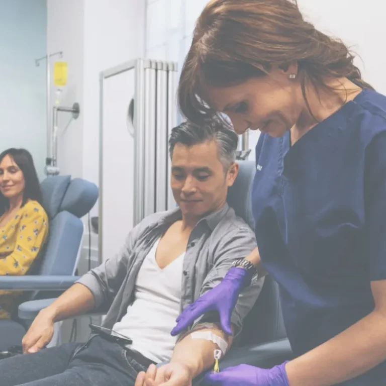 A nurse takes blood from a seated man while another woman waits in the background, both in a medical clinic where IV Booster treatments and antioxidant therapies are offered.