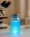 A small glass phial containing a glowing blue liquid, known for the benefits of Methylene Blue, sits on a lab table with a microscope and test tubes blurred in the background.