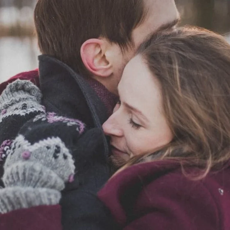 A woman and a man embrace outdoors in winter clothing, with the woman’s eyes closed and knitted mittens visible. Snow and trees are in the background, capturing a moment of winter wellbeing in the crisp, fresh air.