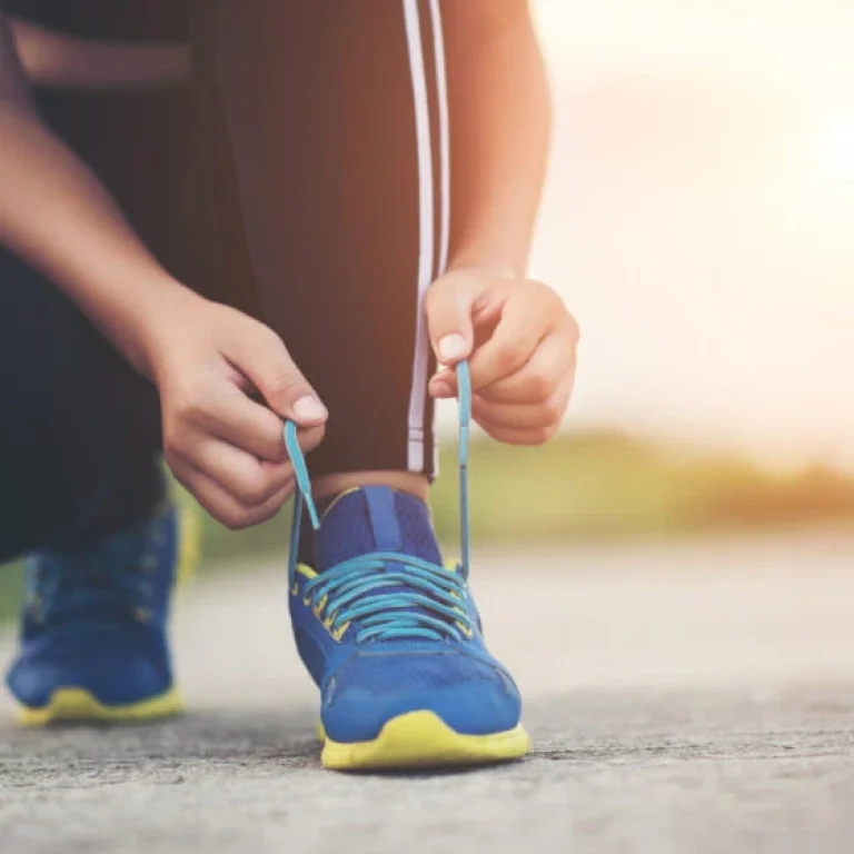Kneeling on the ground and tying the shoelaces of a blue trainer, a person in black trousers with white stripes prepares for peak athletic performance outdoors in natural light.