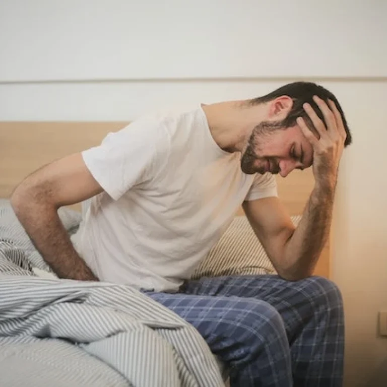 A man in pyjamas sits on the edge of a bed, holding his head with one hand and looking down, appearing unwell—perhaps in need of an instant relief hangover cure.