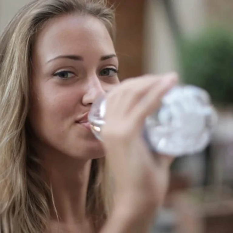 A woman with long blonde hair drinks from a clear plastic water bottle whilst looking at the camera in an outdoor setting, highlighting the importance of dehydration treatment and staying hydrated.