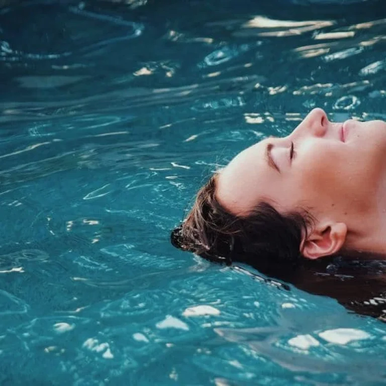 A person floats on their back in a swimming pool with eyes closed, surrounded by clear blue water, enjoying a sense of cleansing and renewal.