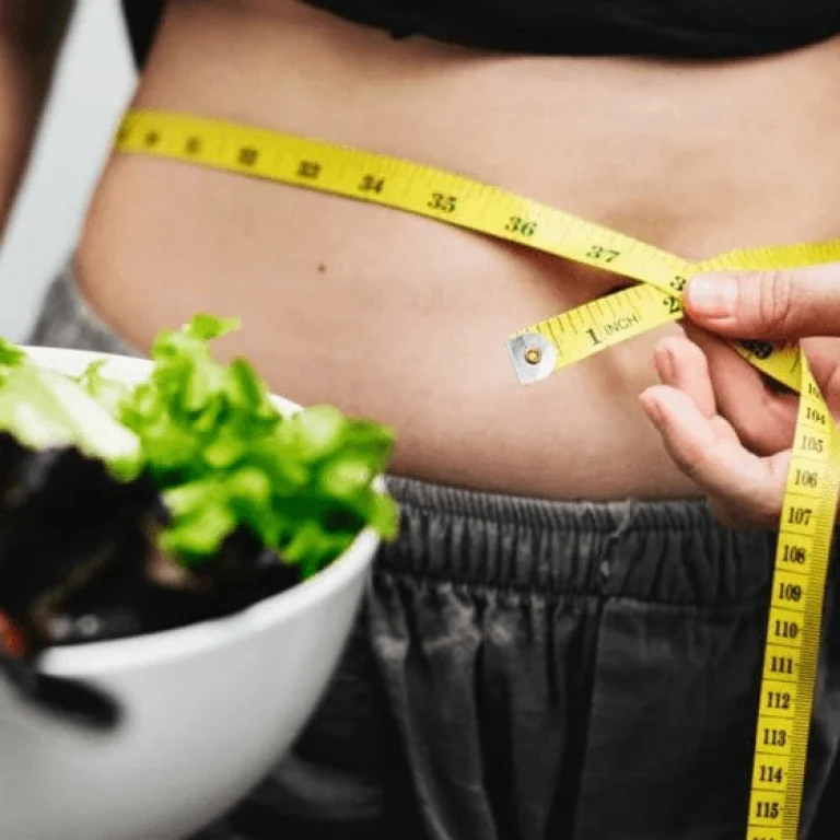 A person measures their waist with a tape measure whilst holding a bowl of salad with lettuce and tomatoes, highlighting their commitment to weight loss.