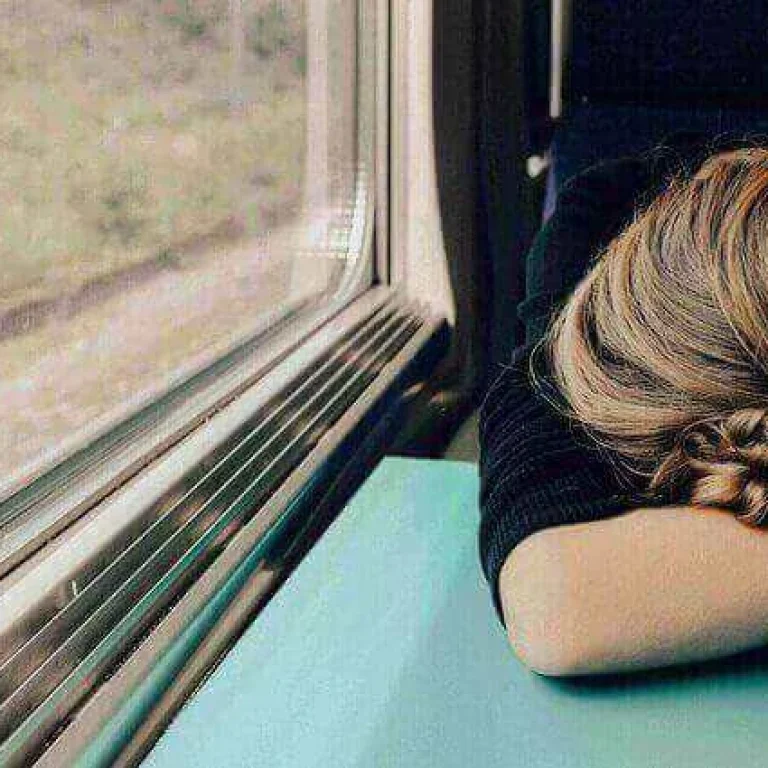 Person with light brown hair rests their head on folded arms on a table next to a window, possibly on a train, quietly pondering what glutathione is and its role in overall wellbeing.