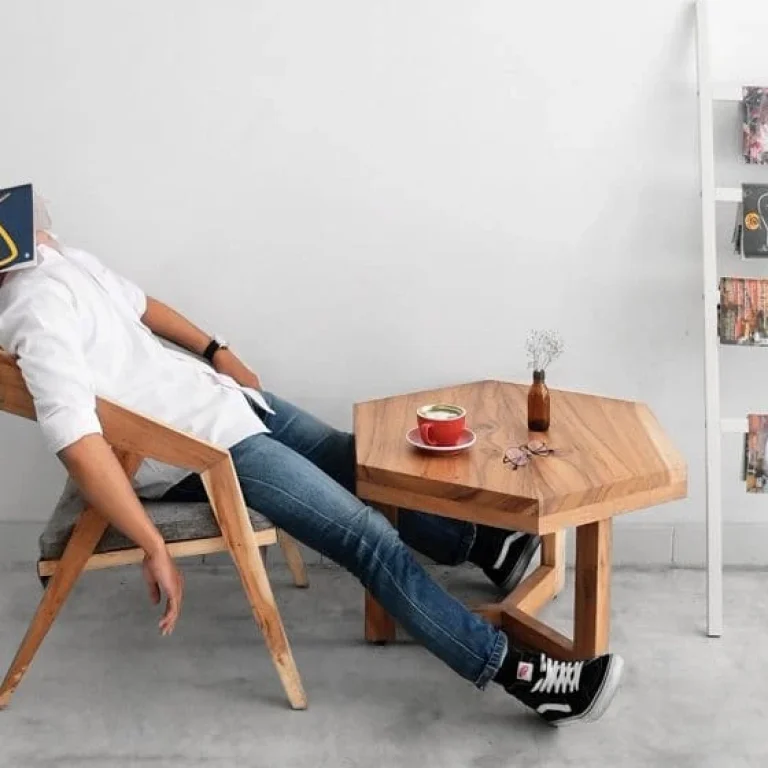 A person sits slouched in a wooden chair with a book covering their face, next to a small table holding a drink and a vase, beside a magazine rack against a white wall.
