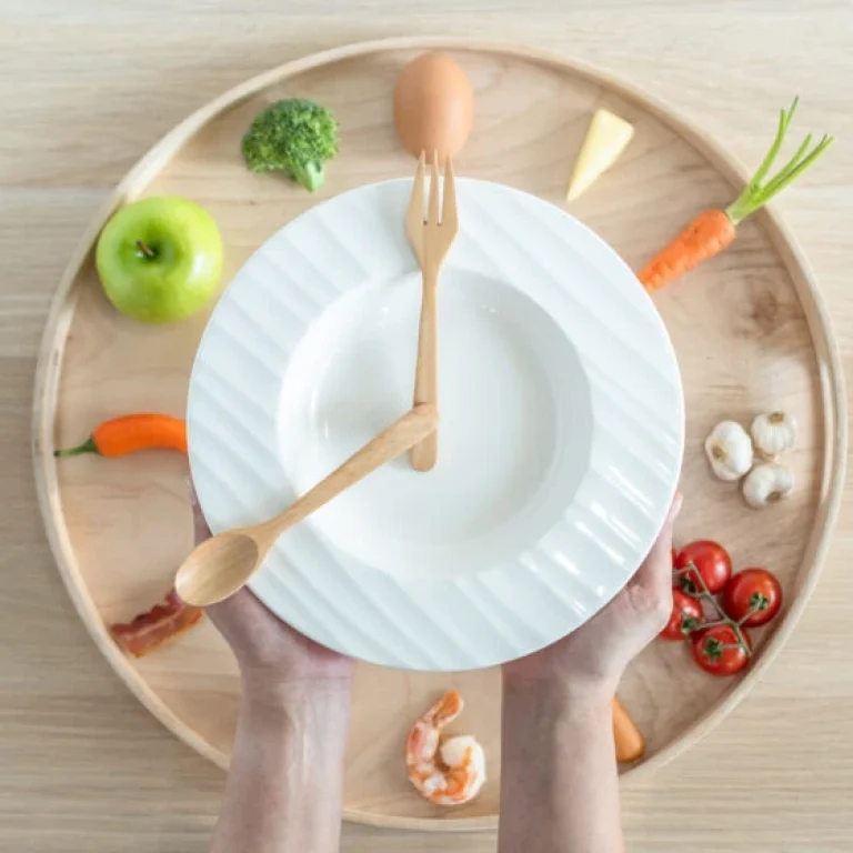 A person holds a white plate with wooden utensils arranged like clock hands, symbolising ways to boost metabolism; the plate rests on a tray with fruit and vegetables arranged in a circle.