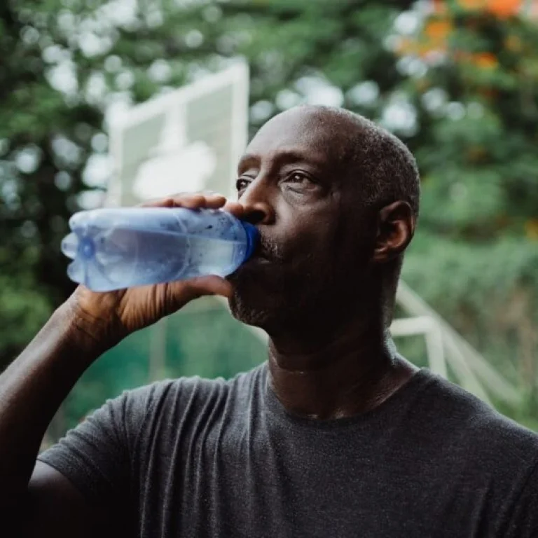 A man in a dark T-shirt drinks water from a blue bottle outdoors, staying hydrated to help combat dehydration and anxiety, with greenery and a basketball hoop blurred in the background.