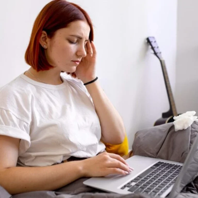 A woman with short red hair sits in bed using a laptop, looking unwell and touching her head, possibly due to Vitamin B12 deficiency, with tissues nearby and a guitar in the background.