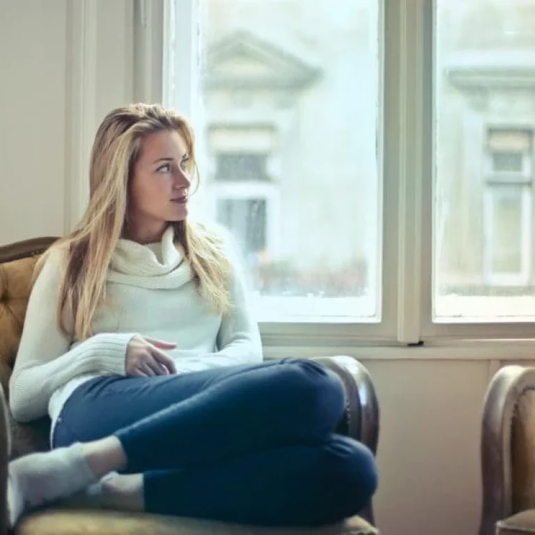 A woman in a white jumper and blue jeans sits cross-legged on an armchair, looking out of a window during lockdown in a well-lit room with vintage decor.