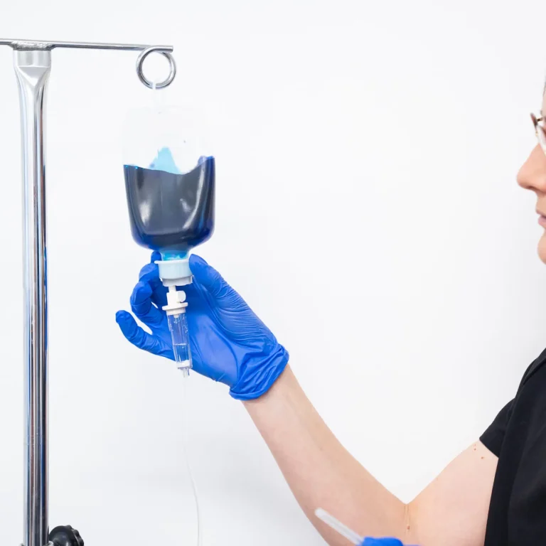 A healthcare worker wearing blue gloves holds a transparent drip bag filled with methylene blue liquid, attached to a drip stand, against a white background.