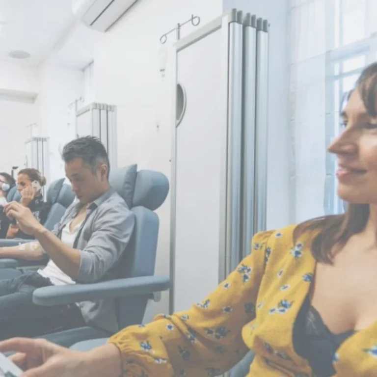 Four adults sit in reclining chairs in a bright, clinical room, each appearing relaxed and engaged in individual activities during their IV therapy sessions.