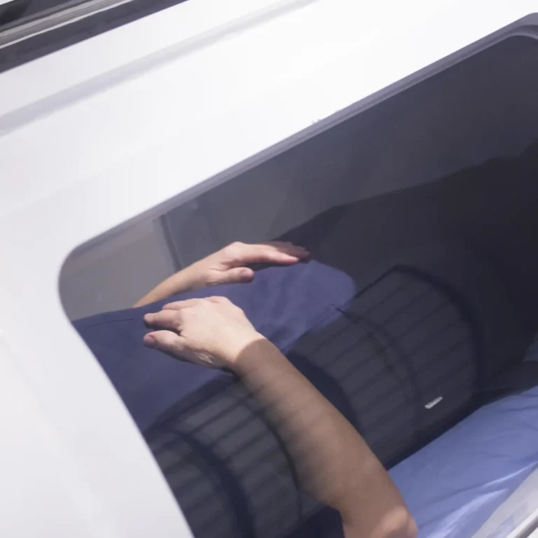 A person lies inside a Hyperbaric Oxygen Therapy machine, with their hands folded on their abdomen and visible through a window.