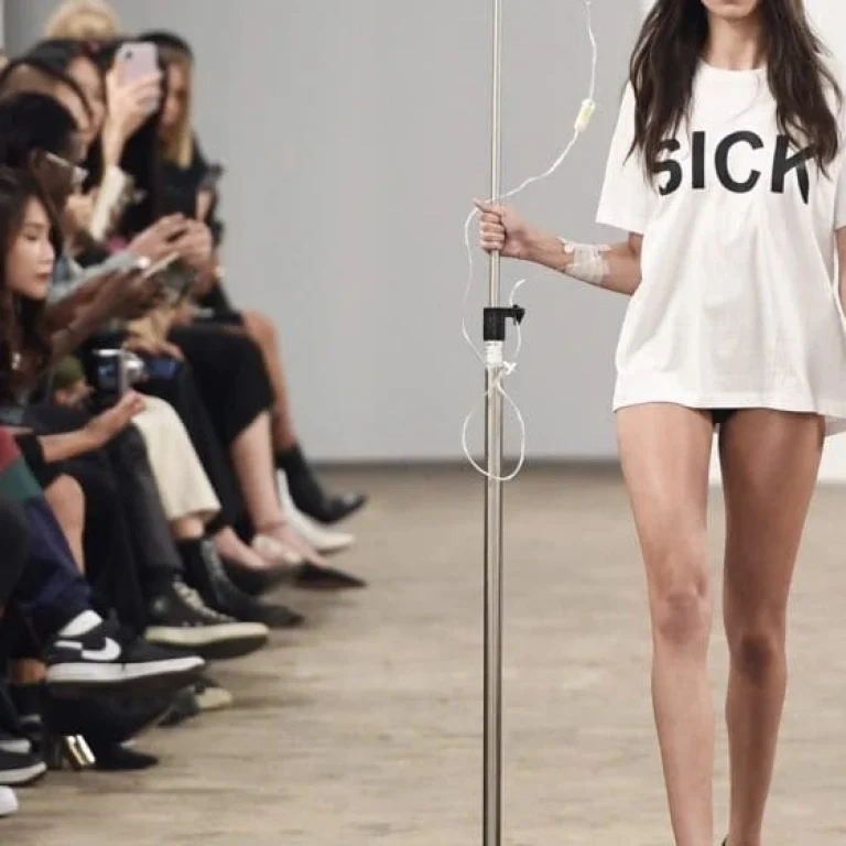 A model walks down a catwalk wearing a white T-shirt with the word SICK printed on it, holding a drip stand—a striking nod to health and IV treatments—while members of the audience watch and take photos.