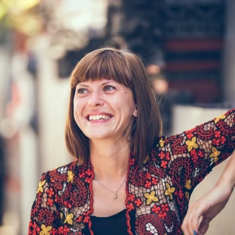 A woman with short brown hair smiles whilst leaning her arm on a wall, wearing a colourful floral-patterned top—radiating vitality that reflects NAD+'s role in anti-ageing and supporting the immune system. The background is softly blurred.