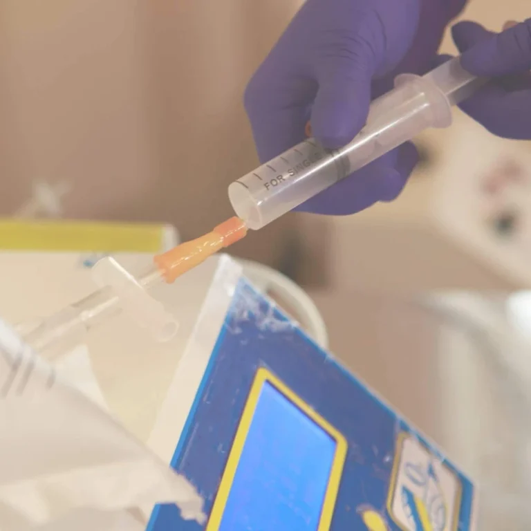 A person wearing purple gloves injects liquid into a medical tube using a syringe near a blue and yellow device, preparing for ozone therapy.