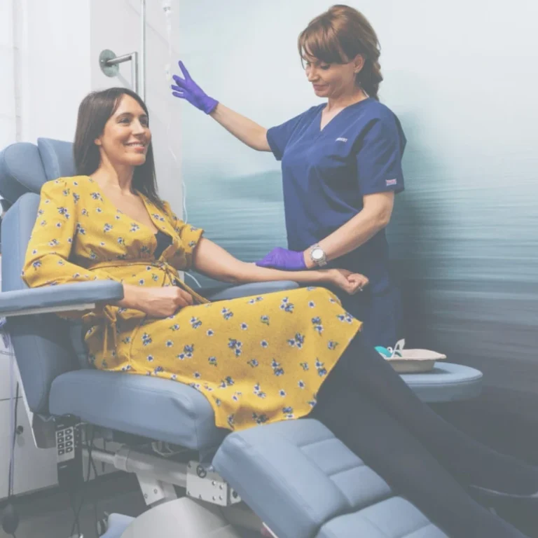 A woman in a yellow dress sits in a medical chair whilst a healthcare professional in scrubs and gloves prepares her for a Skin Brightening IV procedure.