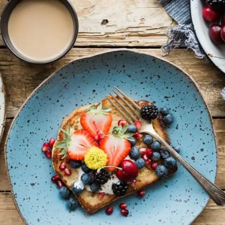 A plate with toast topped with yoghurt, superfoods like strawberries, blueberries, blackberries, pomegranate seeds, and a yellow edible flower sits beside a bowl of cherries and a cup of coffee—perfect to supercharge your healthy new year resolutions.