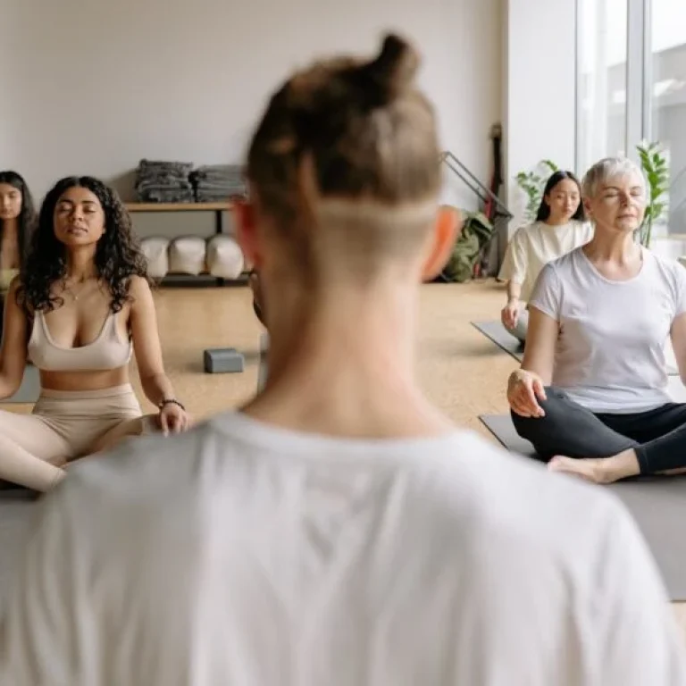 A group of people sit cross-legged on yoga mats in a bright room, meditating with eyes closed, embracing healthy ageing as they follow an instructor in the foreground.