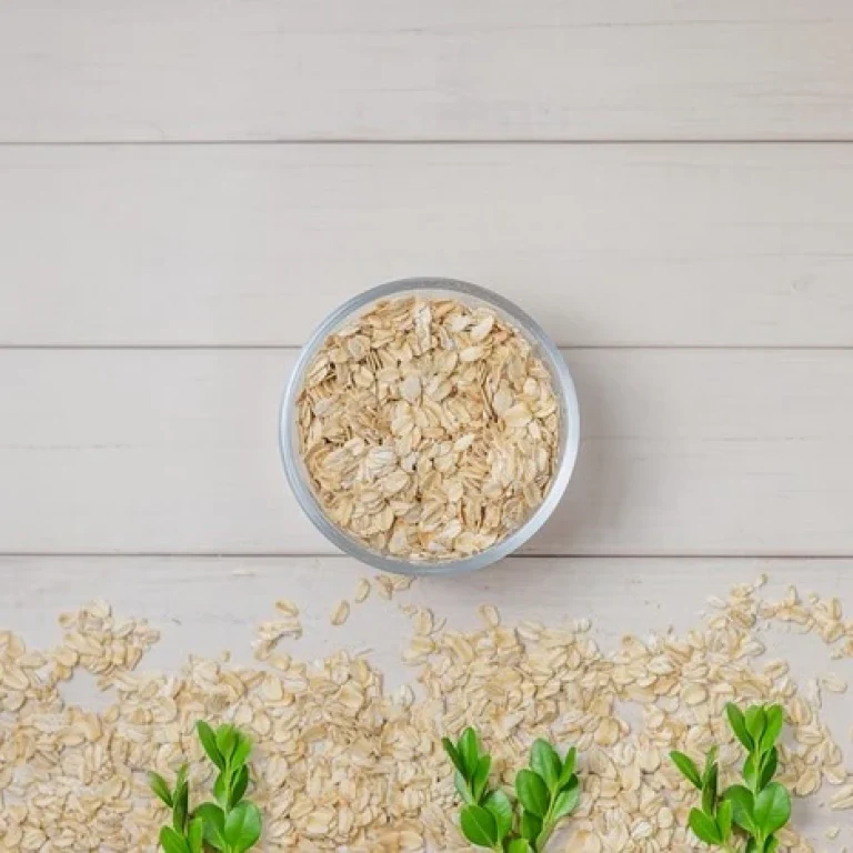 A bowl of porridge oats sits on a light wooden surface, with scattered oats and green leaves arranged at the bottom—highlighting the health benefits and cysteine content found in this nutritious breakfast choice.