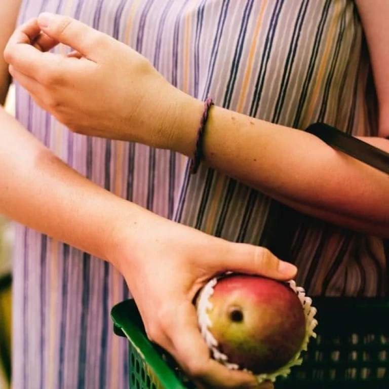 A person in a striped shirt holds a green shopping basket and picks up a mango in a supermarket, mindful of hidden risks that certain foods may pose for those on a restricted diet.