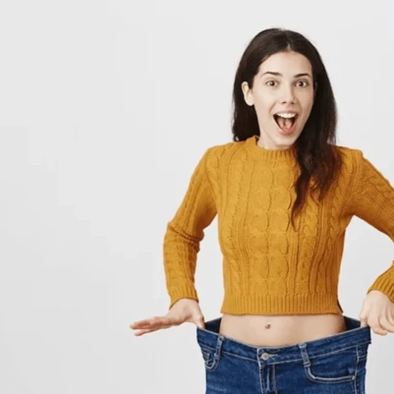 Woman in a mustard jumper holding out oversized jeans, smiling with a surprised expression—showcasing the impressive results of NAD+ Weight Loss against a plain background.