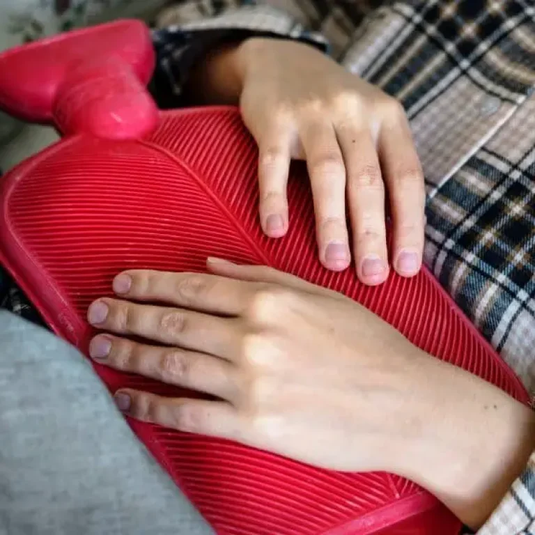 A person lying down holds a red rubber hot water bottle against their stomach with both hands, highlighting common misconceptions about managing Lyme Disease symptoms.