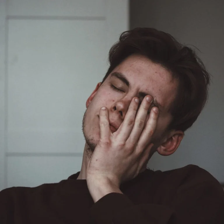 A person with short brown hair rests their face on their hand, eyes closed, appearing tired or stressed—common symptoms of Vitamin B12 deficiency—in an indoor setting with neutral-coloured walls.