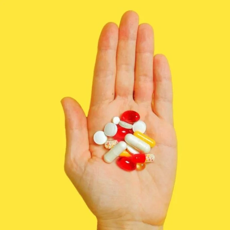 A hand holding various pills and capsules against a solid yellow background, illustrating how vitamin absorption works.