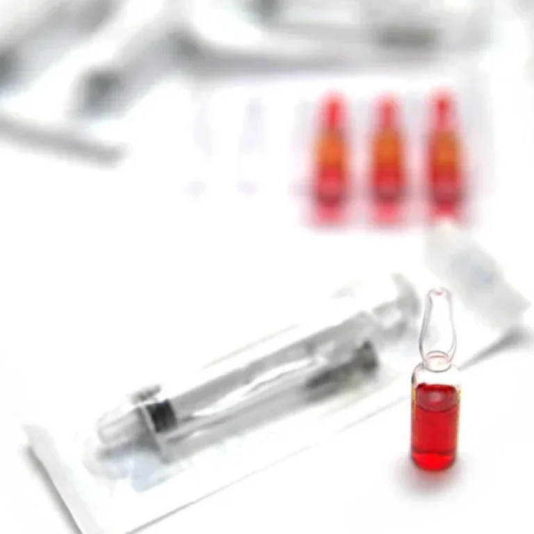 A glass ampoule filled with red liquid stands beside a packaged syringe, hinting at discussions on Vitamin B12 Injections Dosage and Frequency, with more ampoules and syringes blurred in the background.
